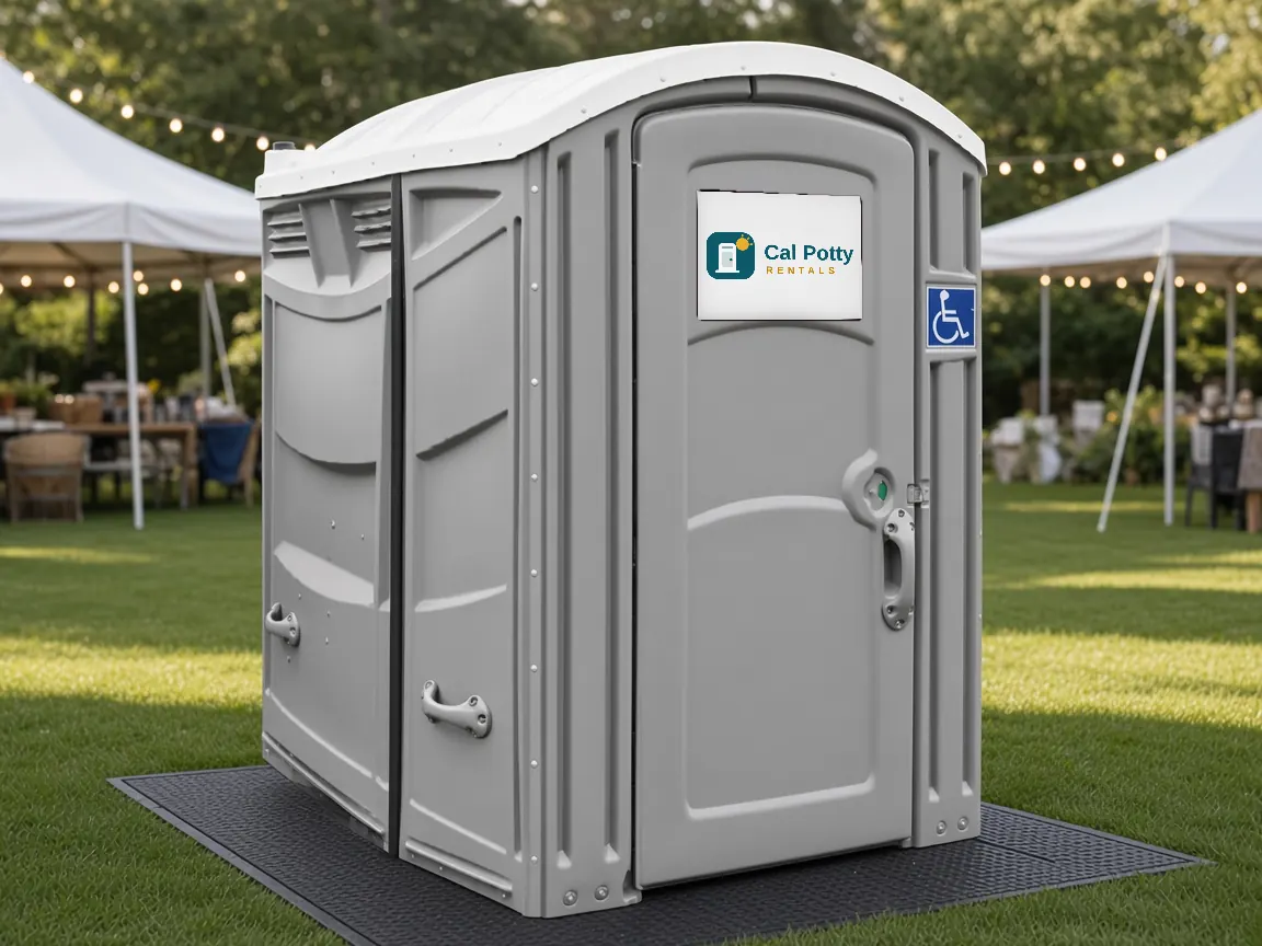 A grey ADA-accessible portable restroom with a wheelchair sign on a rubber mat at an outdoor event under white tents
